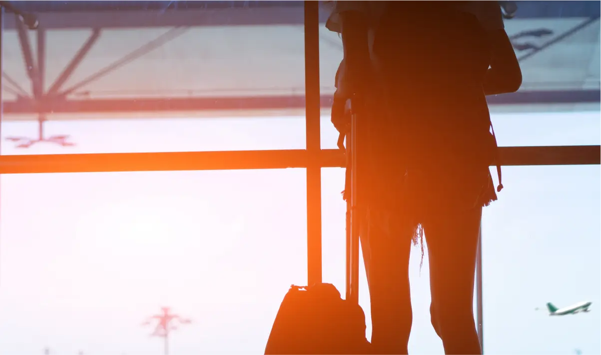 Silhuet af kvinde, der står med bagage i lufthavnsterminal ved solnedgang